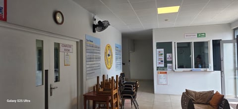 Interior of a medical office waiting room with wooden chairs, information posters on walls, and a reception desk with glass windows