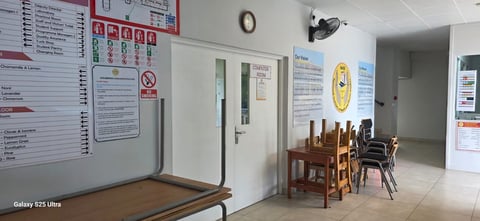 A simple office or break room with wooden furniture, informational posters on walls, and basic décor including a clock and speaker.
