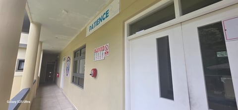 Hospital corridor with yellow walls, Patience sign, glass-fronted offices, and white doors along the hallway