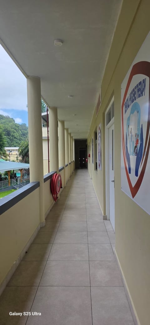 Long covered corridor with cream-colored walls and columns, overlooking a river and green hills, with a red emergency sign visible on the right